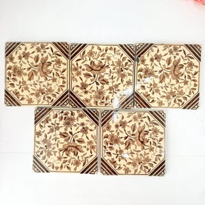 May include: Five square ceramic tiles featuring a cream-colored background with a dark brown floral pattern. Each tile displays a central flower surrounded by smaller blossoms and leaves, framed by a dark brown border with diagonal corner stripes.