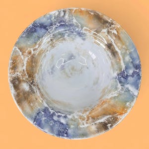 May include: A round ceramic bowl with a marbled design. The bowl features a blend of colours, including white, blue, brown, and gold. The interior is a glossy white, and the rim has a textured, stone-like appearance. The bowl is set against an orange background.