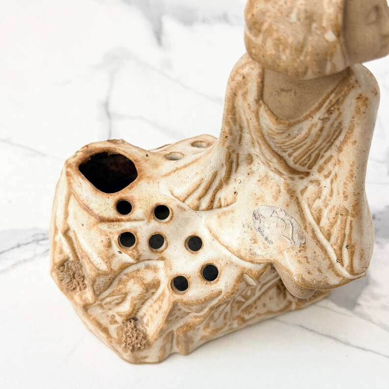 A close-up view of a weathered ceramic Buddha figure, sitting on a textured base, with visible holes for incense smoke. The statue is set against a marble surface, showcasing intricate details of the folds in the robe and the serene expression.