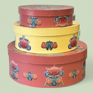 May include: Three round, decorative boxes stacked on top of each other. The largest box is a muted red, the middle box is yellow, and the top box is red. Each box has a repeating floral design in orange, blue, and green. The boxes are nested together.