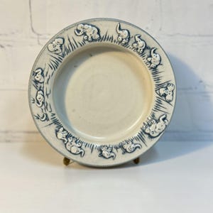 以下が含まれることがあります： 青い縁と青い象のデザインのボーダーが付いた、クリーム色のセラミックプレート。 プレートは丸く、金色のスタンドに置かれています。 象はさまざまなポーズをとっています。