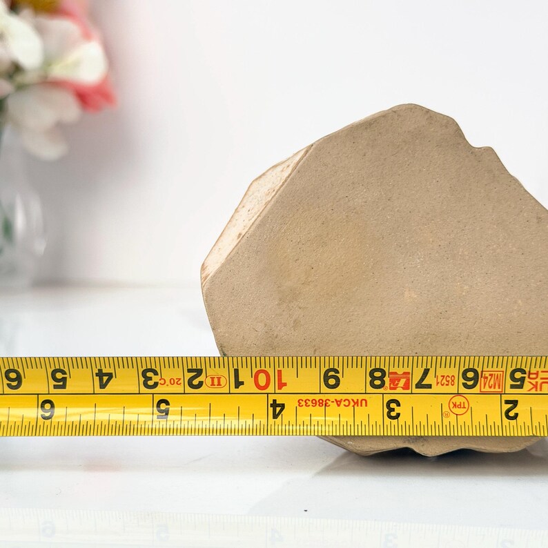 A yellow measuring tape marked from 1 to 10 inches lies across a large, irregularly shaped clay object on a white surface, with a vase of flowers in the background.
