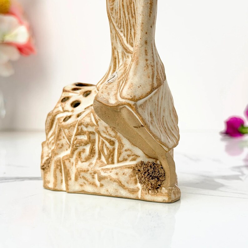 Close-up of a beige ceramic statue with carved patterns on a white marble surface; blurred flowers in the background.
