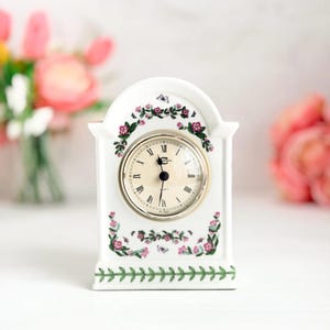 May include: A white ceramic mantel clock with a round clock face and Roman numerals. The clock is decorated with pink floral and green leaf patterns. The clock face is surrounded by a silver-coloured metal ring.