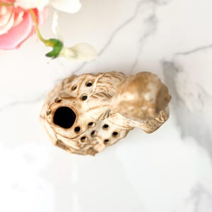 Overhead view of a small, beige, turtle-shaped ceramic ocarina with multiple finger holes, resting on a white marble surface near delicate pink and white artificial flowers.