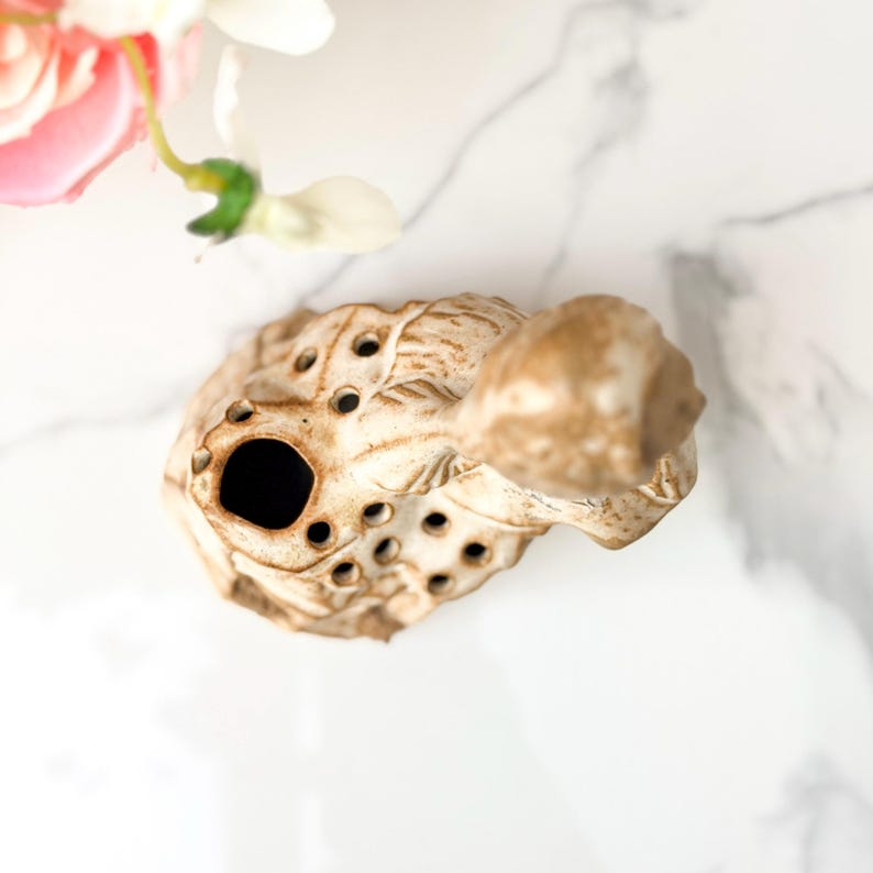 Overhead view of a small, beige, turtle-shaped ceramic ocarina with multiple finger holes, resting on a white marble surface near delicate pink and white artificial flowers.