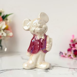Iridescent ceramic Mickey Mouse figurine in a red vest stands on a marble surface, with blurred flowers in the background.