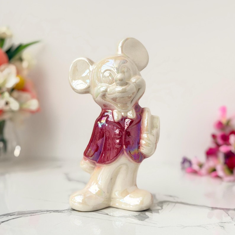 Iridescent ceramic Mickey Mouse figurine in a red vest stands on a marble surface, with blurred flowers in the background.