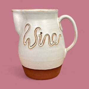 Puede incluir: Jarra de cerámica blanca con base y asa marrones. La palabra "Wine" está inscrita en cursiva en la parte delantera. La jarra está sobre un fondo rosa.