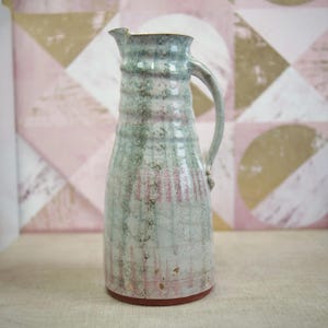 Op de afbeelding: Een handgemaakte keramische kan met een lange, slanke vorm en een gebogen handvat. De kan heeft een getextureerd oppervlak met verticale lijnen en een gevlekt patroon van groen, roze en wit. De basis is roodbruin.