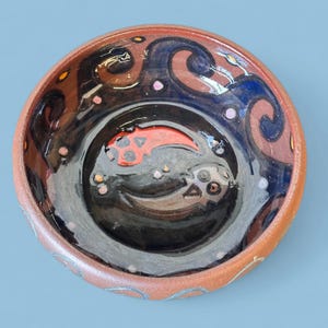 May include: A handmade ceramic bowl with a dark interior and a reddish-brown exterior. The bowl features a unique design with swirling patterns in brown and blue, and a red fish-like shape in the center. Small, colorful dots add detail.