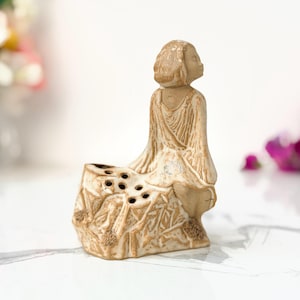 Pode incluir: Uma escultura de cerâmica bege apresentando uma figura sentada sobre uma base texturizada com múltiplas aberturas circulares. A figura tem uma cabeça lisa e arredondada e uma vestimenta drapeada com linhas gravadas visíveis.