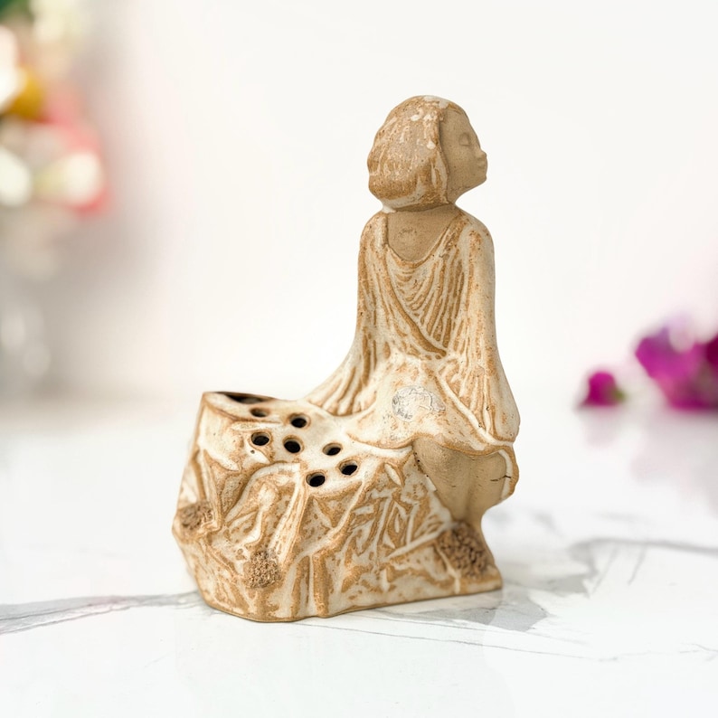 A ceramic figure of a person sitting in a meditative pose on a small, textured rock, with holes at the base, set against a white marble surface.