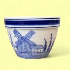 May include: White ceramic plant pot featuring a blue windmill design. The pot has a blue band around the rim and a detailed illustration of a Dutch windmill scene. The design includes a windmill, houses, and stylized plants.