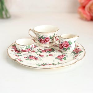 Könnte beinhalten: Ein Miniatur-Teeservice aus Porzellan bestehend aus einem runden Tablett, einer kleinen Teetasse, einem Milchkännchen und einer winzigen Schale. Jedes Teil ist mit rosa Rosen und grünen Blättern auf weißem Grund sowie Goldrändern verziert.