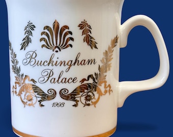 Puede incluir: Taza de cerámica blanca con ribete dorado y un diseño dorado con las palabras "Buckingham Palace" y el año "1993". La taza tiene un asa curva y una banda dorada alrededor de la base.