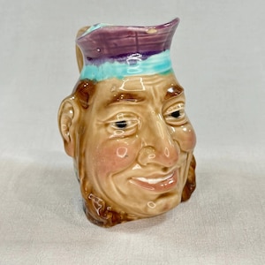 May include: A vintage ceramic pitcher shaped like a smiling man's face. The pitcher features a tan face with rosy cheeks, brown hair, and a purple and turquoise hat. The handle is on the left side, and the pitcher has a wide, open mouth.