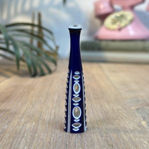 May include: A tall, cobalt blue glass vase with a narrow neck and a decorative pattern of oval and diamond shapes. The vase is approximately 8 inches tall and sits on a wooden surface. The top of the vase is clear.