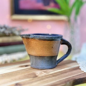 Puede incluir: Una taza de cerámica blanca con un asa negra y el texto "But First, Coffee" en negrita negra. La taza está llena de un líquido oscuro, probablemente café, y se encuentra sobre una superficie de color claro.