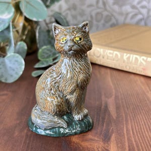 May include: A ceramic cat figurine in a seated pose. The cat has a brown and tan coat with yellow eyes and is perched on a green base. The figurine is detailed with textured fur and is set against a blurred background.