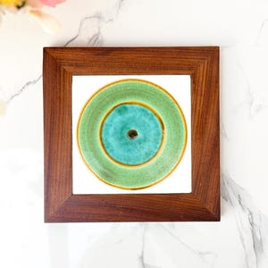 May include: A square wooden frame containing a white ceramic tile. The tile features a circular design with a central dark brown dot, surrounded by a vibrant turquoise inner ring and a wider, textured light green outer ring with brown edges.