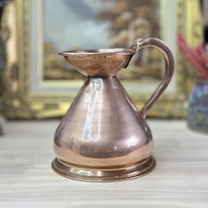 May include: A copper-coloured, vintage-style jug with a handle. The jug has a conical body and a flared top. The handle is curved and attached to the side of the jug. The jug is sitting on a light-coloured surface.