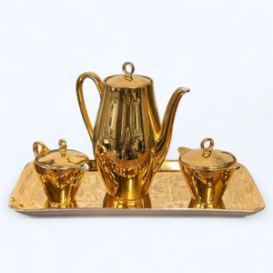 May include: A gold-plated tea set comprising a teapot, creamer, sugar bowl, and serving tray. The set has a highly reflective surface. The teapot features a curved handle and spout. The sugar bowl and creamer have lids with small handles.