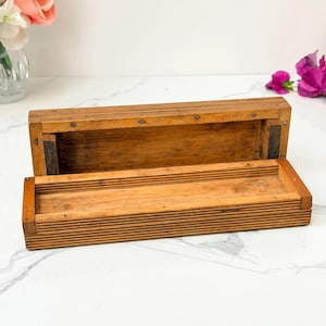 May include: A rectangular wooden box with a hinged lid, featuring a natural light brown finish and a ribbed, textured exterior surface. The box is open, revealing a smooth, hollow interior compartment with visible joinery and small metal nails.