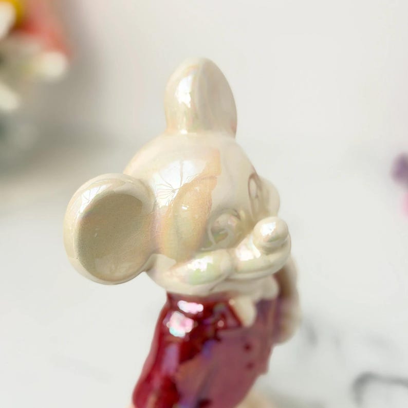 Close-up of a pearlescent, vintage ceramic Mickey Mouse figurine with a red vest, set against a blurred background.