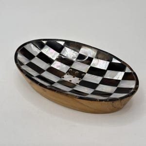 May include: Oval soap dish with a black and white checkerboard pattern. The dish is made of wood and mother-of-pearl. The dish has a light brown wooden base and a decorative design.