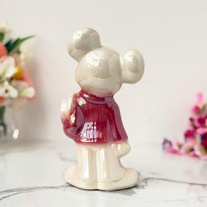 A vintage, pearlescent ceramic mouse figurine wearing a burgundy jacket and cream pants stands on a marble surface, with blurred floral arrangements in the background.