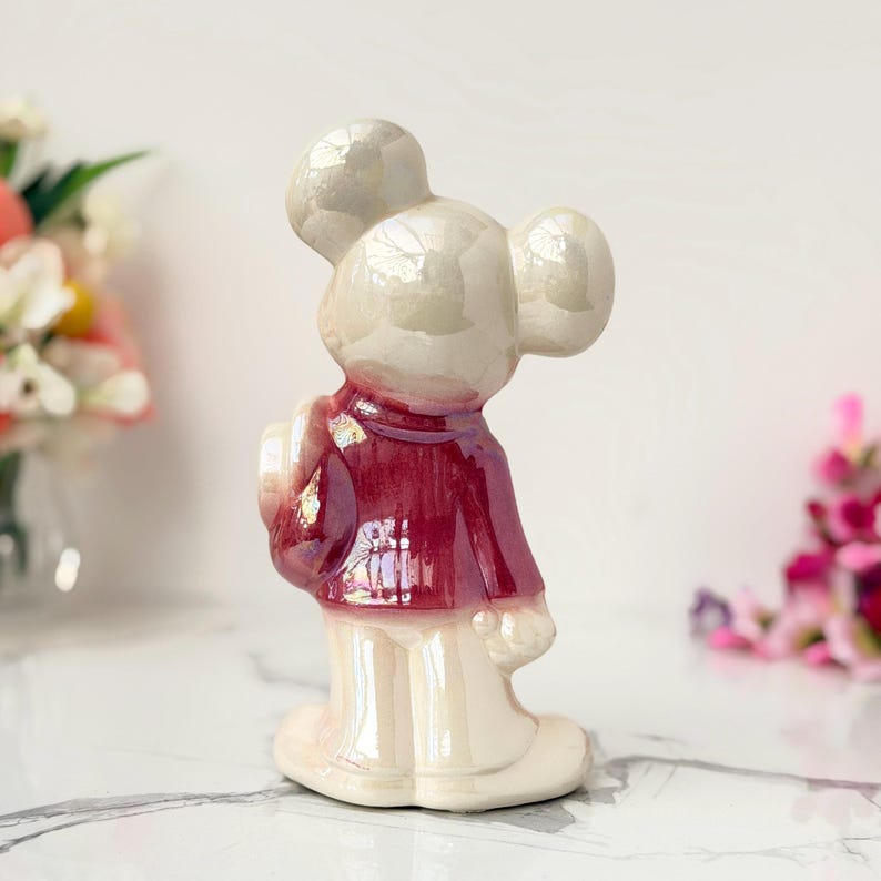 A vintage, pearlescent ceramic mouse figurine wearing a burgundy jacket and cream pants stands on a marble surface, with blurred floral arrangements in the background.