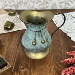 May include: A vintage-style pitcher with a weathered turquoise finish and brass accents. The pitcher features a curved handle and decorative rope detailing with brass ball ornaments. The base and rim are made of brass.