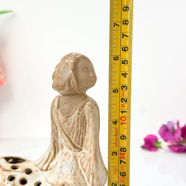 A close-up of a beige ceramic figurine of a seated woman, with her hands folded in her lap, next to a yellow measuring tape, measuring 38 centimeters tall, against a white background with scattered pink and purple flowers.