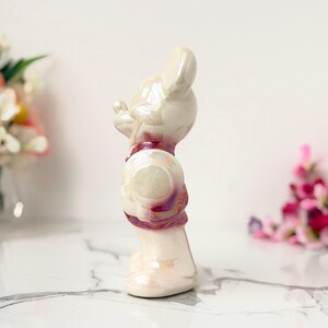 A vintage, iridescent ceramic figurine of a mouse wearing a red scarf stands on a marble surface, with blurred floral arrangements in the background.