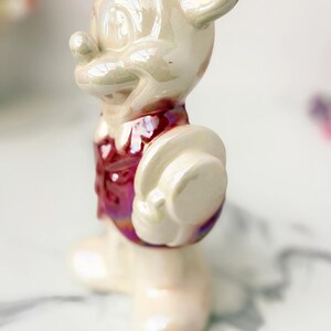 Close-up of a vintage, iridescent ceramic Mickey Mouse figurine wearing a red vest and holding a white hat. The figurine has a pearlescent sheen and sits on a marbled surface.