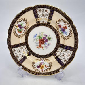May include: A decorative plate with a cream-colored background, featuring a central floral design and surrounding floral and geometric patterns. The plate has a scalloped edge and is accented with gold and navy blue details.