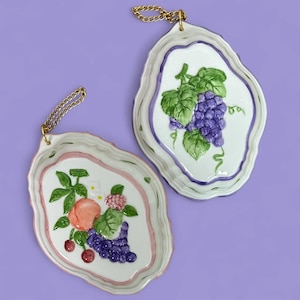 May include: Two ceramic wall plaques with gold chain hangers. One depicts a bunch of purple grapes with green leaves, the other has a peach, cherries, and grapes. Both have scalloped edges and are painted with pastel colours.