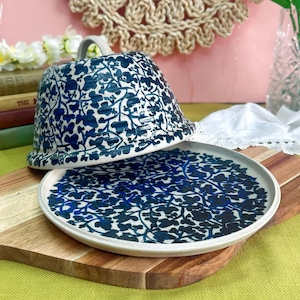 May include: A ceramic cloche and plate set with a blue floral pattern. The cloche has a handle and sits atop a matching plate. The design features a dark blue floral motif against a white background. The set is displayed on a wooden chopping board.