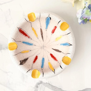 Könnte beinhalten: Ein runder, cremefarbener Aschenbecher mit strukturierter Oberfläche. Er zeigt ein farbenfrohes, stilisiertes Design aus strahlenförmigen Formen in Rot, Blau, Gelb und Braun. Der Aschenbecher hat vier gelbe Zigarettenablagen am Rand.
