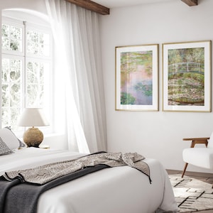 May include: Two framed landscape paintings of water lilies and a bridge over a pond. The paintings are in gold frames and are hanging on a white wall. The paintings are in a bedroom setting with a bed and a chair.