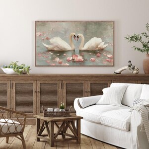 May include: A framed artwork depicts two white swans forming a heart shape with their necks, floating on water with pink flower petals. The artwork has a wooden frame and is displayed above a wooden cabinet.