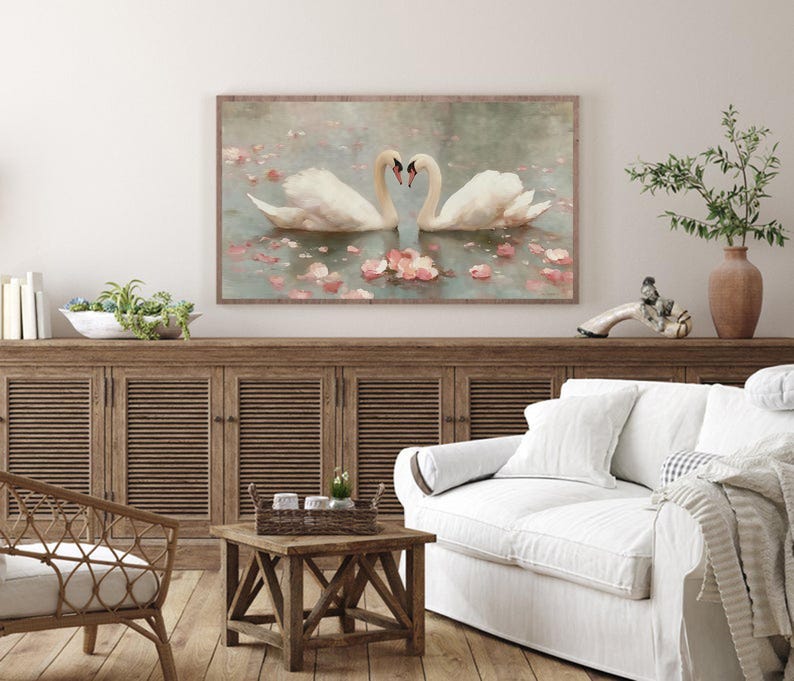 May include: A framed artwork depicts two white swans forming a heart shape with their necks, floating on water with pink flower petals. The artwork has a wooden frame and is displayed above a wooden cabinet.