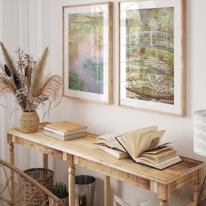 May include: Two framed prints of water lily paintings by Claude Monet. The prints are in a light wood frame and are hanging on a white wall above a wooden console table. The table is decorated with books, a vase of dried flowers, and a wicker basket.