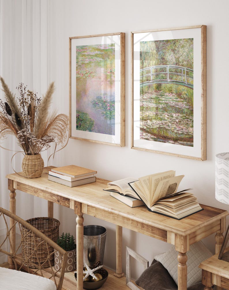 May include: Two framed prints of water lily paintings by Claude Monet. The prints are in a light wood frame and are hanging on a white wall above a wooden console table. The table is decorated with books, a vase of dried flowers, and a wicker basket.