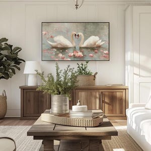 May include: A living room scene featuring a large framed artwork of two swans forming a heart shape. The room includes a white sofa, wooden furniture, a coffee table with a woven tray, and a potted plant in a woven basket. The walls are white.