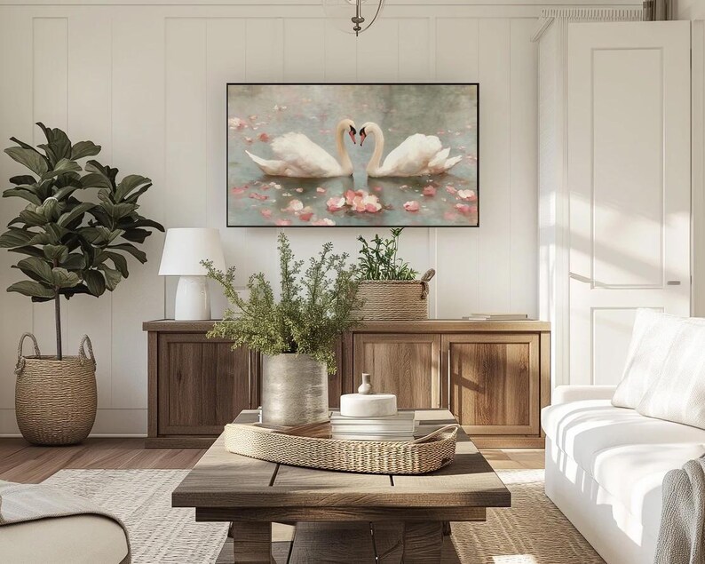 May include: A living room scene featuring a large framed artwork of two swans forming a heart shape. The room includes a white sofa, wooden furniture, a coffee table with a woven tray, and a potted plant in a woven basket. The walls are white.