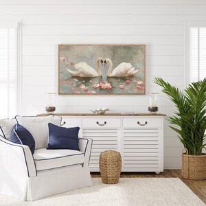 May include: A framed artwork depicts two white swans forming a heart shape with their necks, floating in water with pink flower petals. The artwork hangs above a white cabinet with a wooden top, a white armchair with navy blue pillows, and a potted plant.