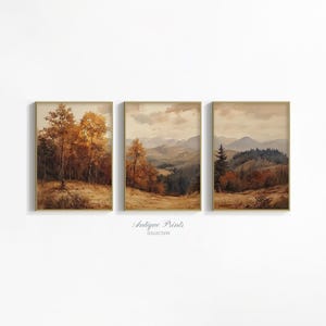 May include: A set of three rectangular framed wall art prints featuring watercolour landscapes in shades of brown, gold, and muted green. The images depict autumn forests, rolling hills, and distant mountains under a cloudy sky with text below.