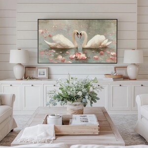 May include: A living room scene featuring a large framed artwork of two swans forming a heart shape with their necks. The artwork hangs above a white cabinet with two matching lamps. A coffee table with a floral arrangement and two white sofas complete the room.